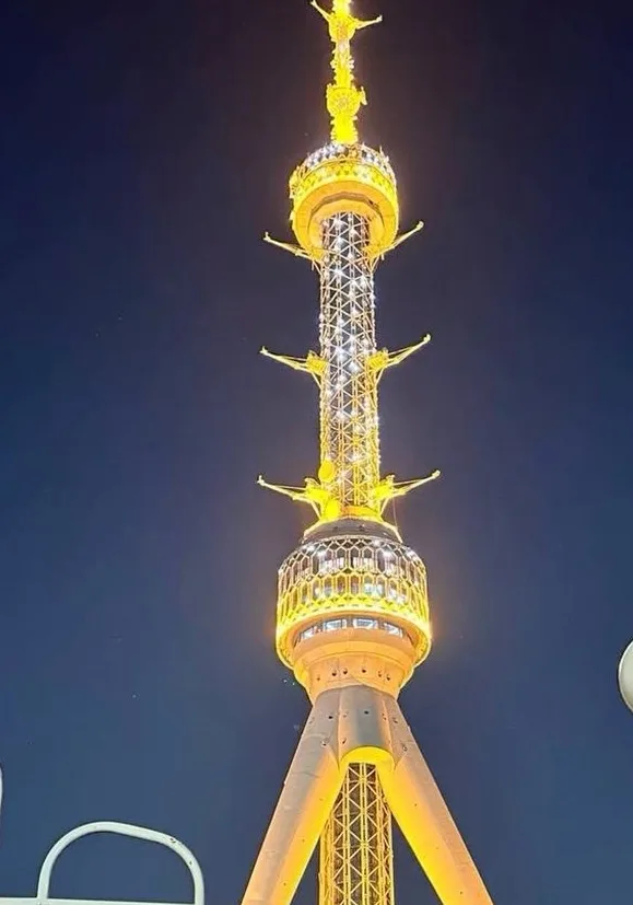 A nighttime photo of a tall, illuminated golden tower with multiple tiers and statues of figures with outstretched arms, set…