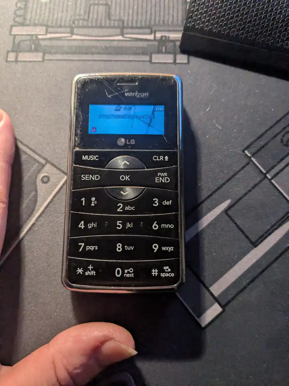 A close-up photo shows a person's fingers holding an old, scratched LG flip phone with a Verizon logo, its small blue screen…