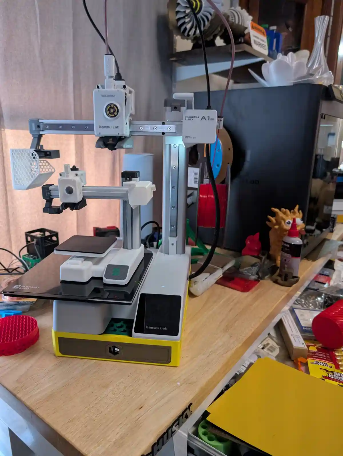 A Bambu Lab A1 3D printer sits on a cluttered wooden workbench, surrounded by spools of filament, printed parts, and tools…