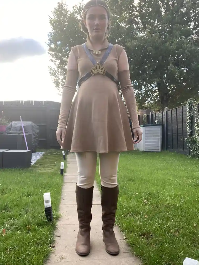 A person stands outdoors on a garden path wearing a brown dress, beige leggings, and brown boots, with a costume harness and…