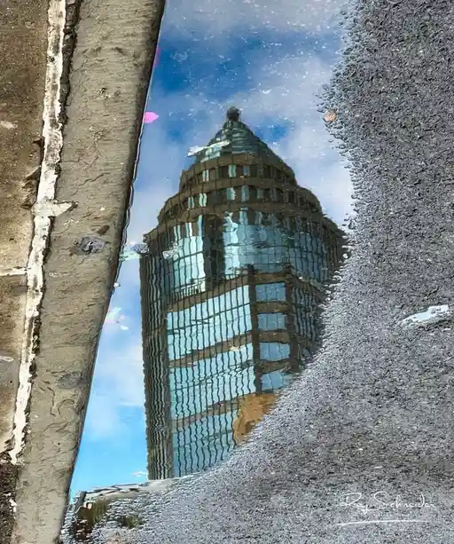 A puddle on asphalt reflects a tall glass skyscraper against a blue sky with clouds. The reflection is distorted by ripples…