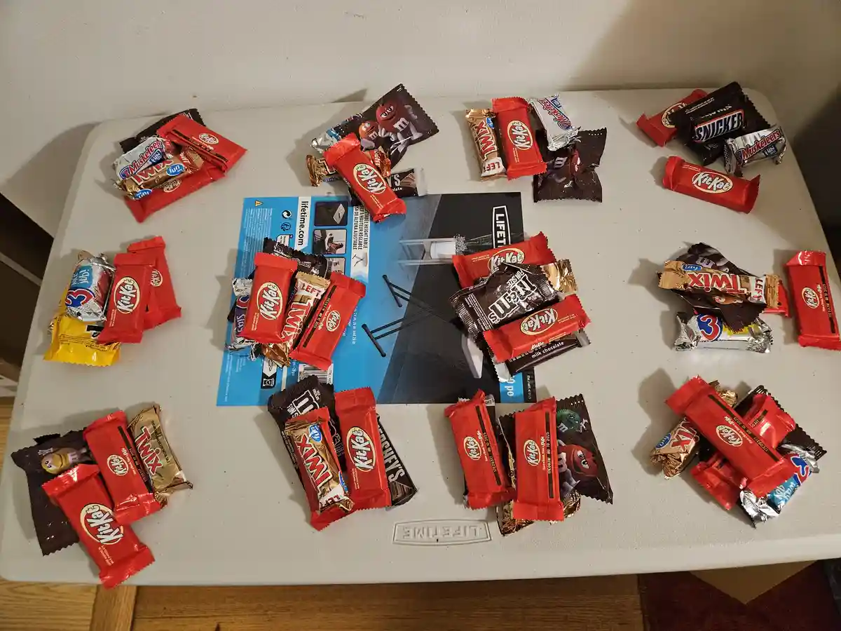 A white folding table is covered with numerous chocolate candy bars, primarily Kit Kats, arranged in piles around a blue…