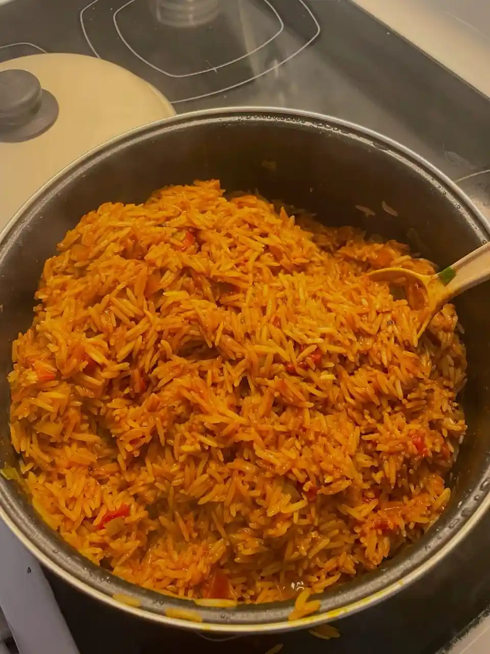 A close-up photo shows a large metal pot filled with vibrant orange jollof rice, with a wooden spoon resting inside. The…