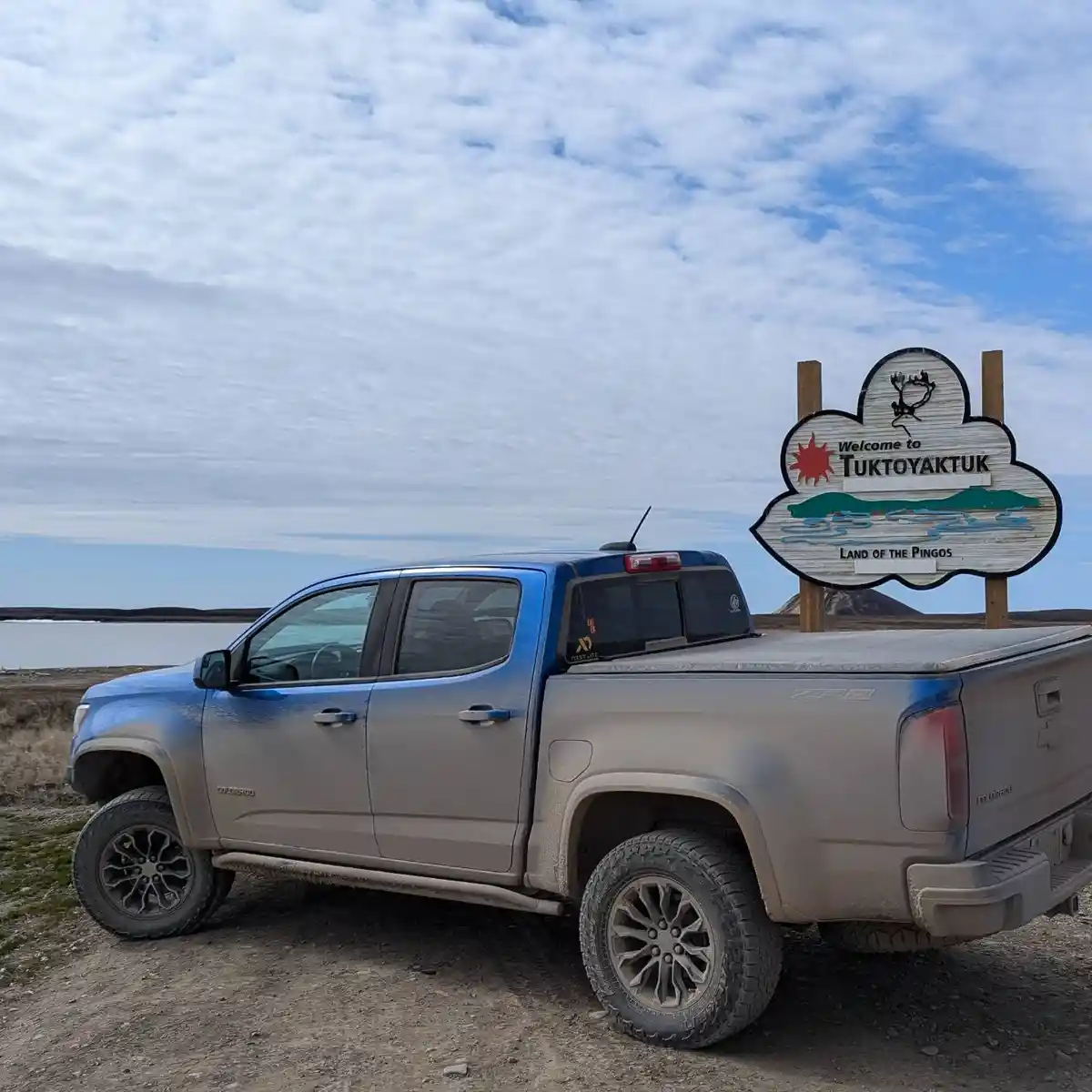 A dusty blue Chevrolet Colorado ZR2 pickup truck is parked beside a wooden welcome sign for Tuktoyaktuk, Nunavut, Canada…