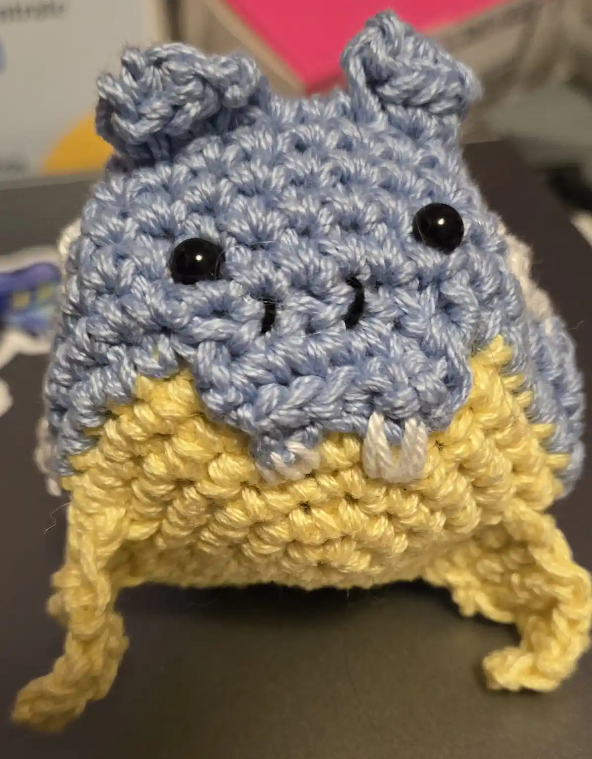 A close-up photo of a handmade crocheted creature with a blue head, black bead eyes, a stitched smile, and white crochet…