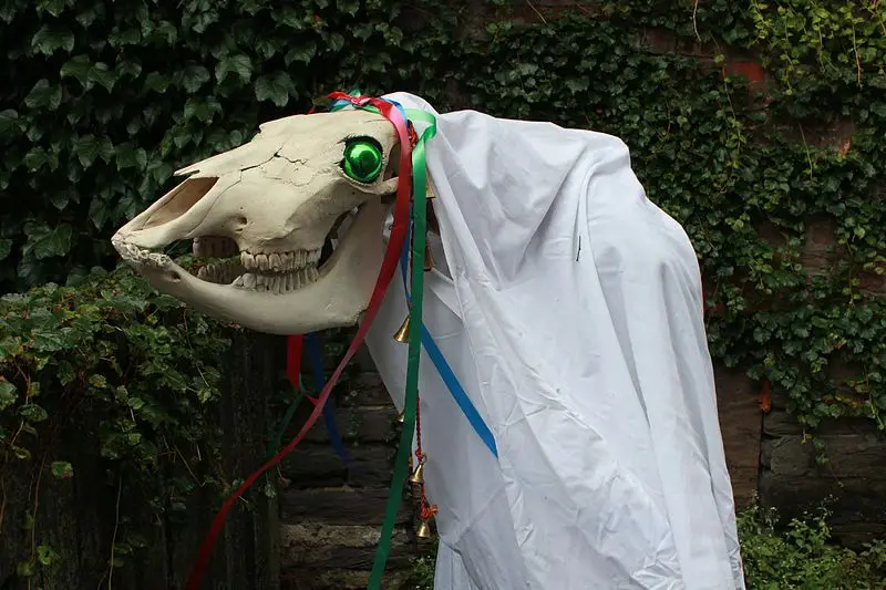 A horse skull with a glowing green eye, draped in a white sheet like a ghost, adorned with colorful ribbons and small bells…
