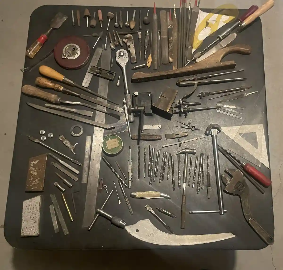 An overhead shot displays a large, dark workbench covered with a vast, meticulously arranged collection of vintage and…