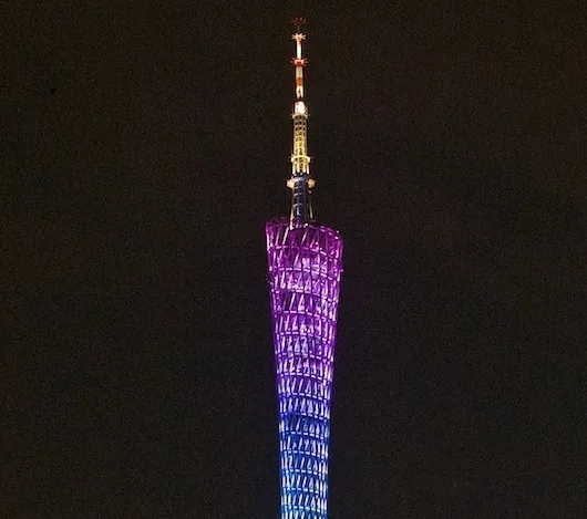 A tall, slender tower illuminated at night with purple and blue lights, tapering to a spire with red and white lights at the…