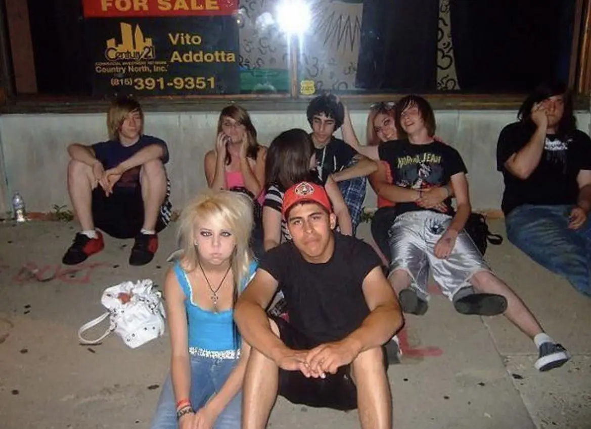 A group of young people, mostly teens, are sitting on a sidewalk at night in front of a 'FOR SALE' sign for a property owned…