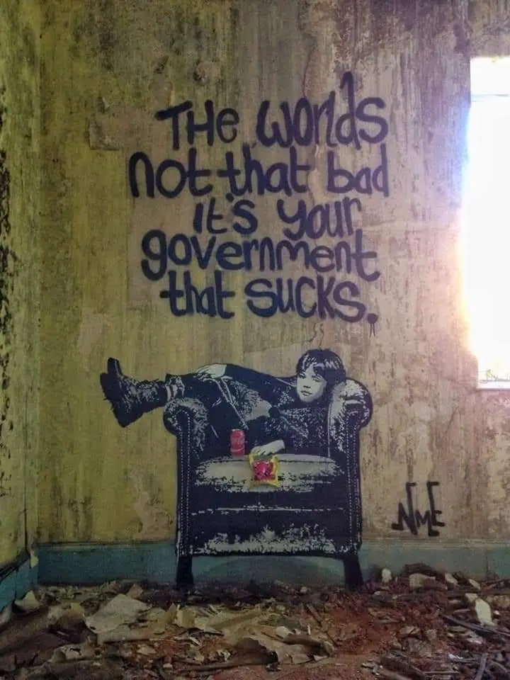 A graffiti mural on a dilapidated, peeling yellow wall inside a ruined building shows a boy lounging in an armchair with a…