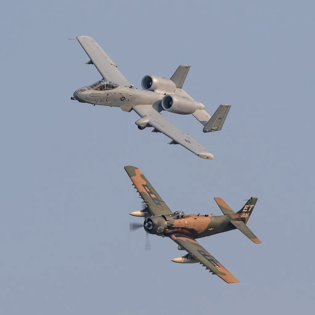 Two military aircraft fly side-by-side against a clear blue sky: a modern twin-engine A-10 Thunderbolt II (Warthog) above…