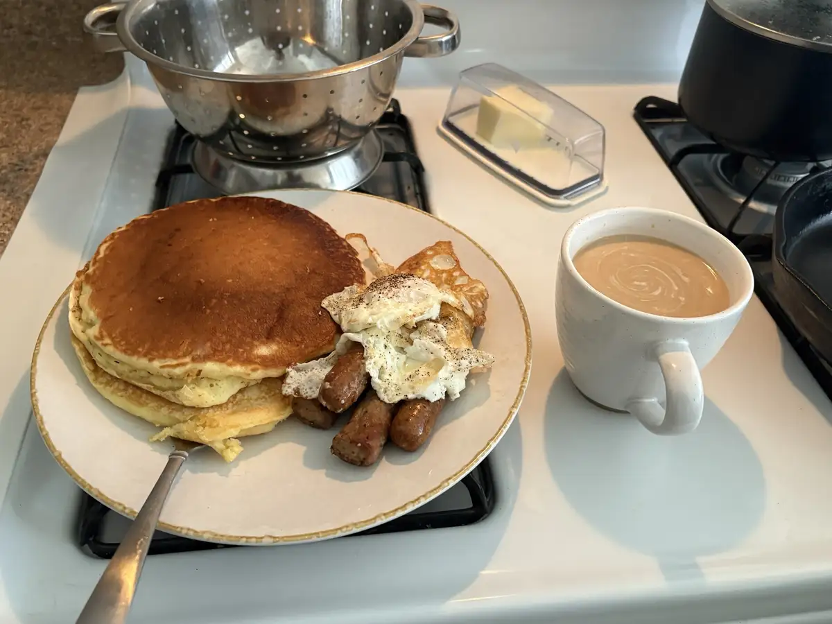 A plate of breakfast food sits on a stovetop, featuring a stack of golden-brown pancakes, fried sausage links, and a…