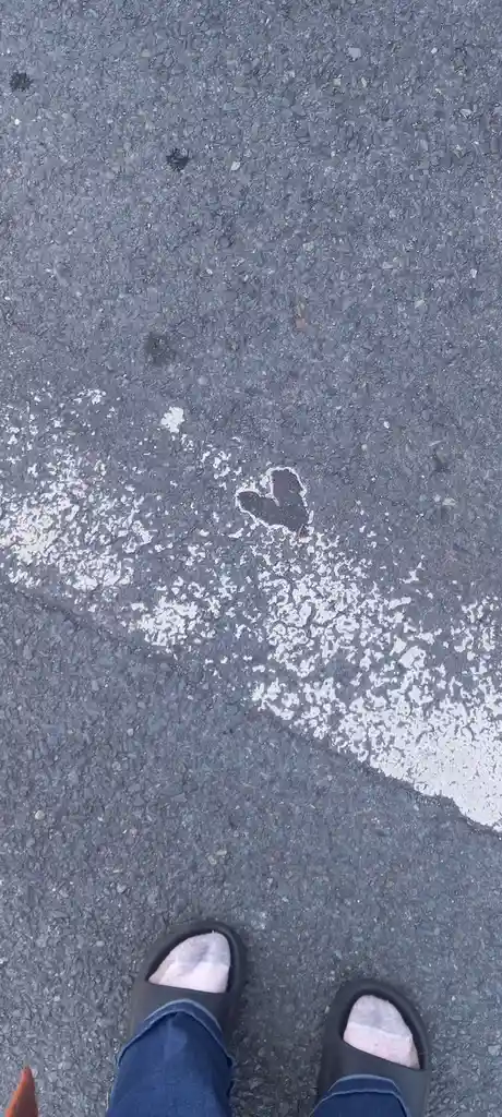 A top-down view shows a person's feet in black sandals and blue jeans standing on a gray asphalt road. A small, distinct…
