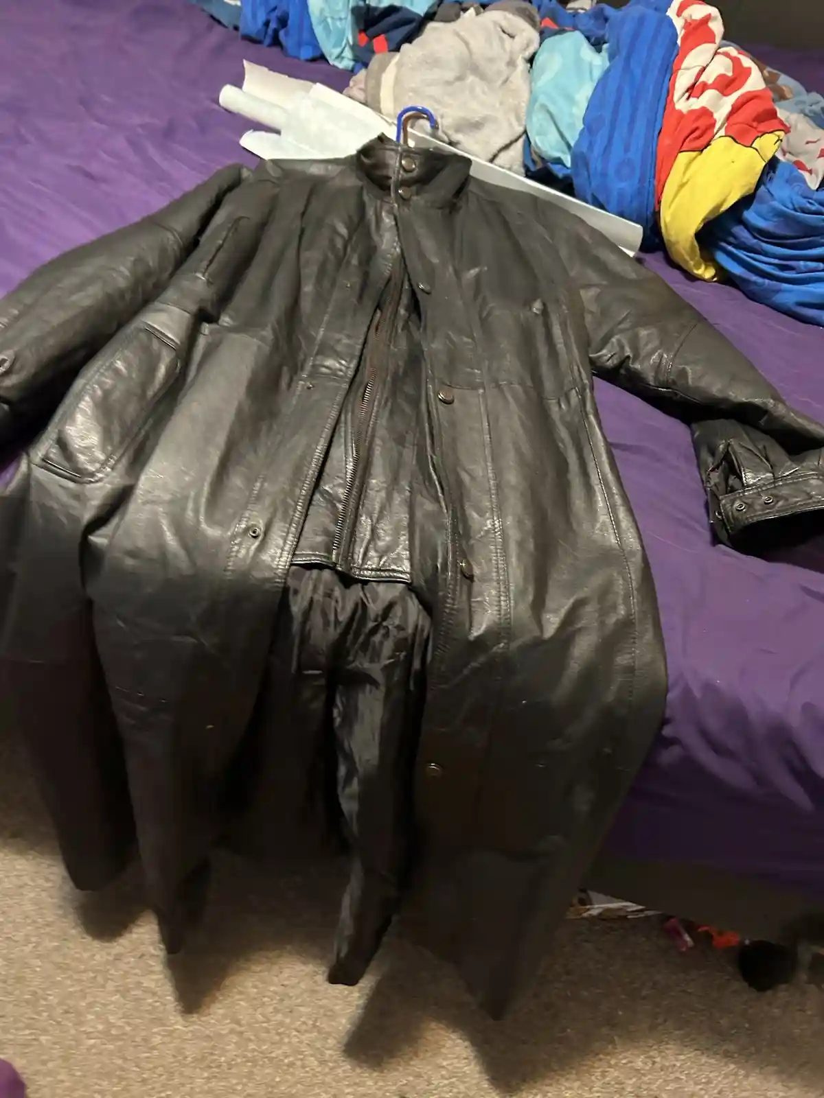 A black leather jacket hangs on a hanger over a purple bedspread, with its bottom half pulled down to reveal a black pair of…