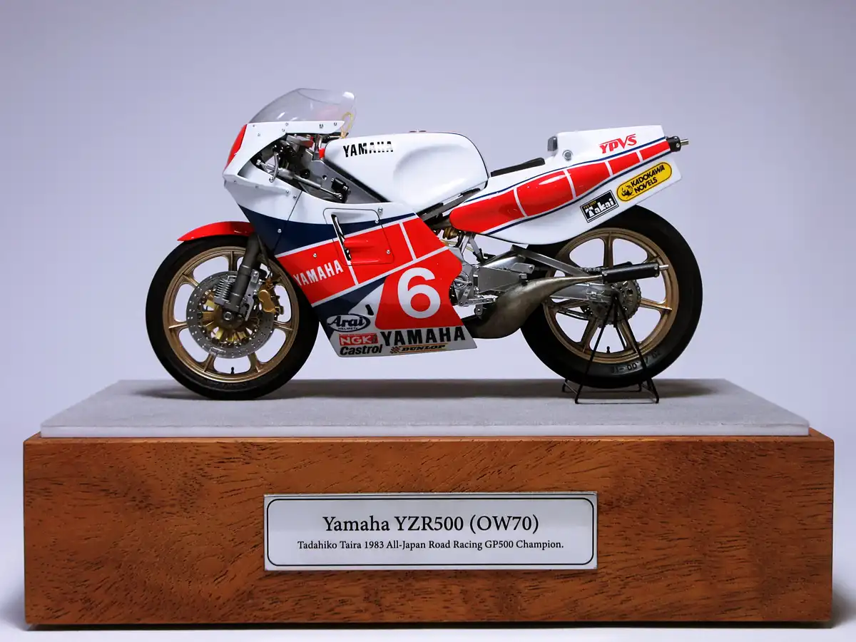 A detailed scale model of a white, red, and blue Yamaha YZR500 racing motorcycle, number 6, displayed on a wooden pedestal…