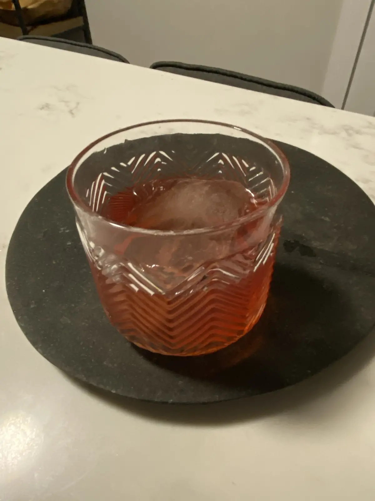 A close-up photo shows a patterned glass containing a reddish-brown liquid and a large ice cube, resting on a black coaster…