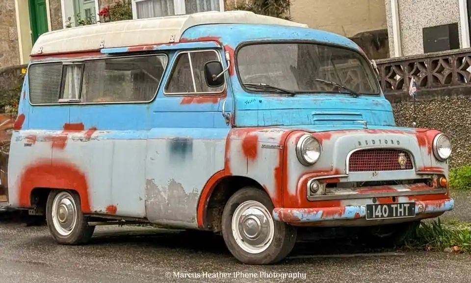 A weathered, rusty Bedford van with peeling blue and red paint is parked on a street. The license plate reads 'I40 THT'. The…