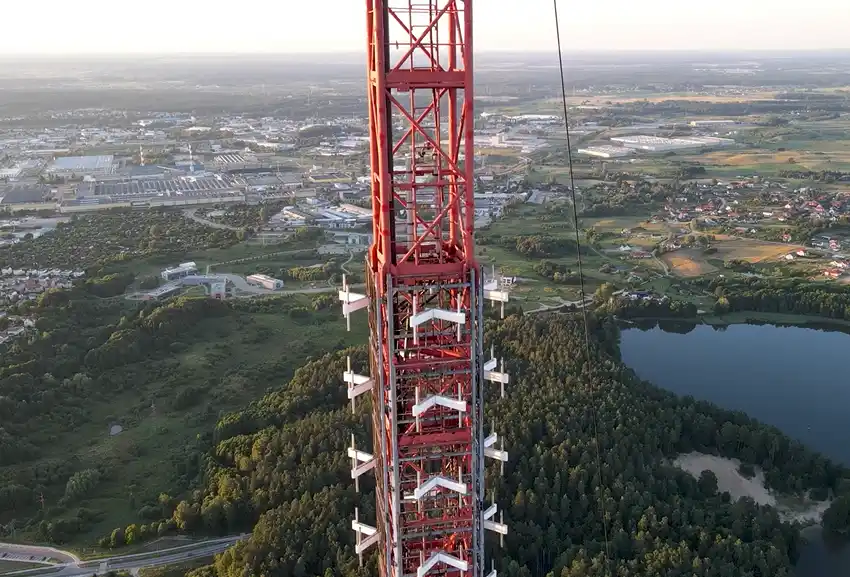 An aerial view shows a tall red lattice tower under construction, surrounded by green forests, a lake, and distant towns…