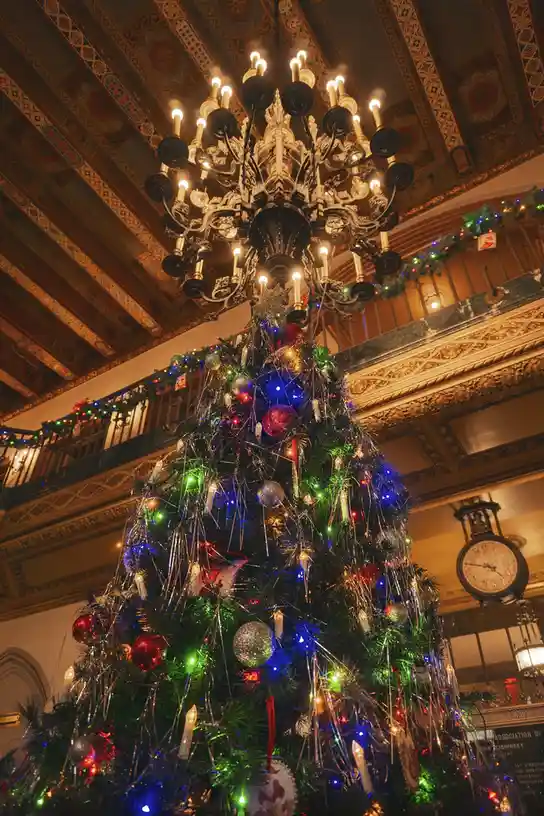 a lavishly decorated christmas tree in an ornate, multi-level building with a large chandelier overhead and a clock visible…
