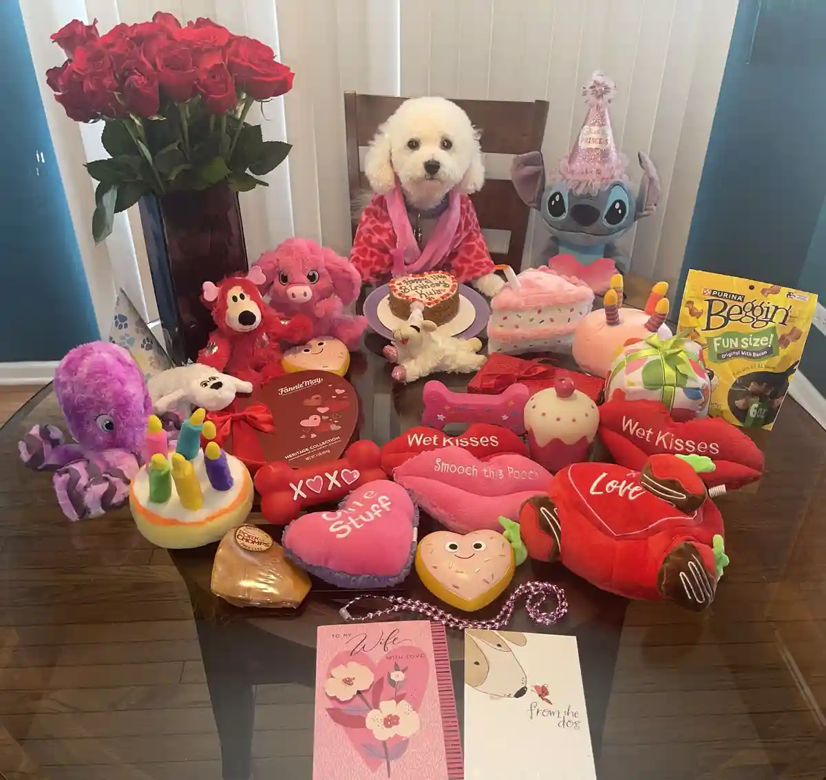 A small white dog wearing a pink robe sits at a glass table surrounded by an extravagant, pink-themed birthday spread for a…