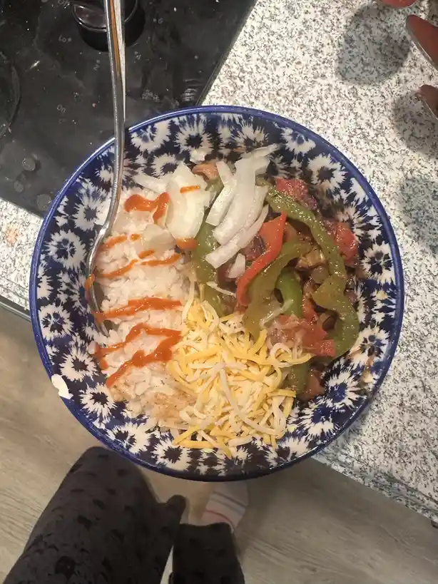 A top-down photo of a blue and white patterned bowl filled with rice, sautéed peppers and onions, shredded cheese, and a…