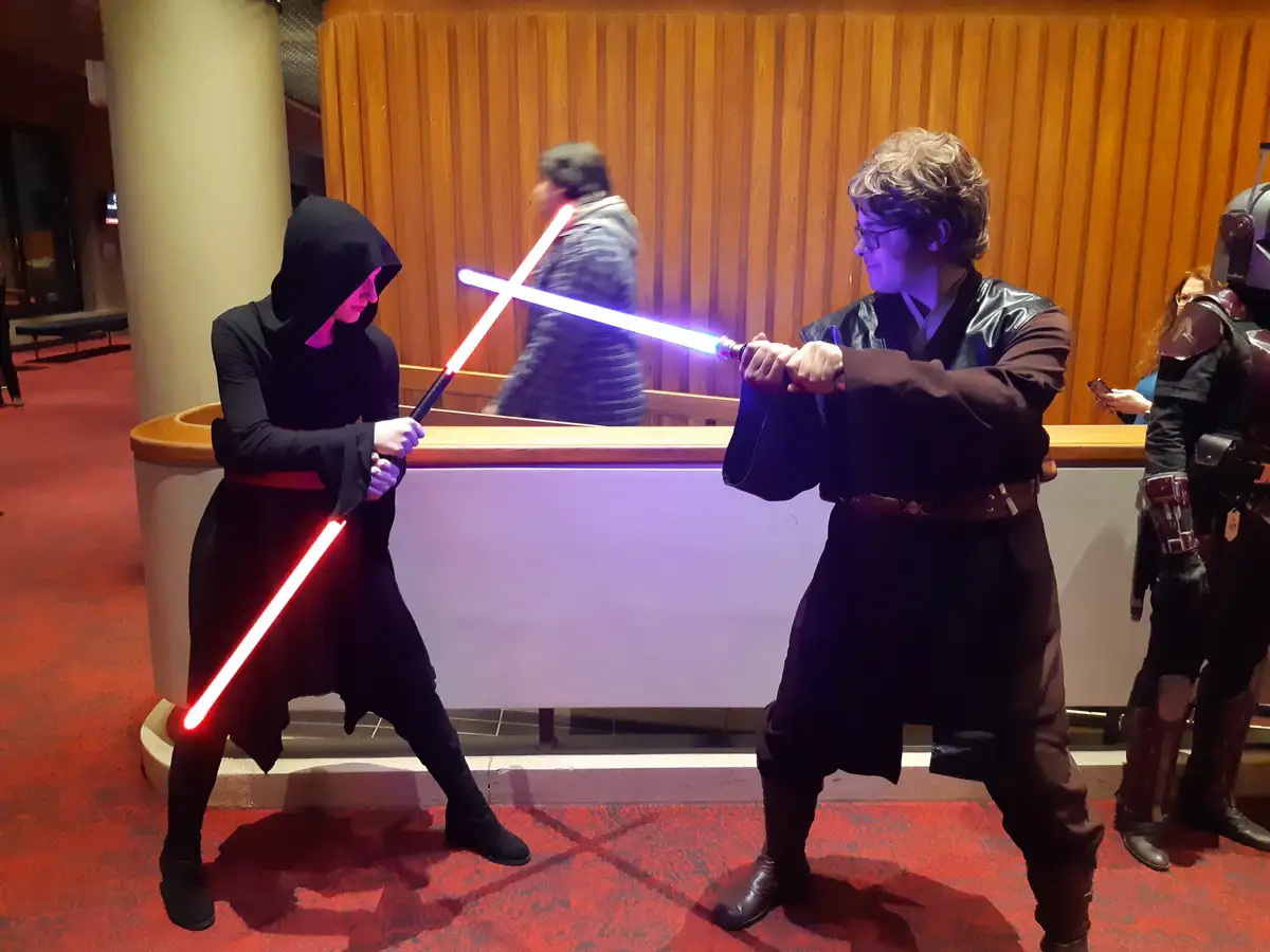 Two people in Star Wars cosplay are engaged in a lightsaber duel indoors, one in black hooded robes wielding a red…