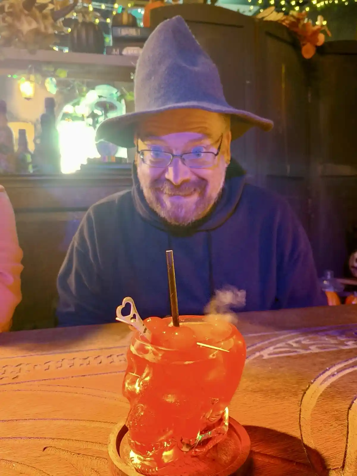 A man with glasses and a blue wizard hat smiles at the camera while sitting at a bar, behind a skull-shaped glass filled…
