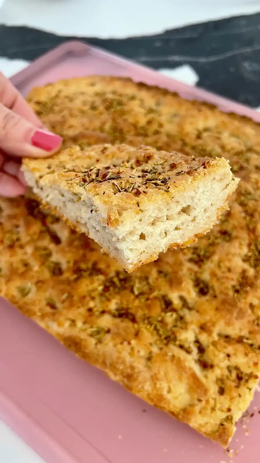 A hand with pink nail polish lifts a slice of golden-brown herb bread from a larger loaf resting on a pink cutting board…