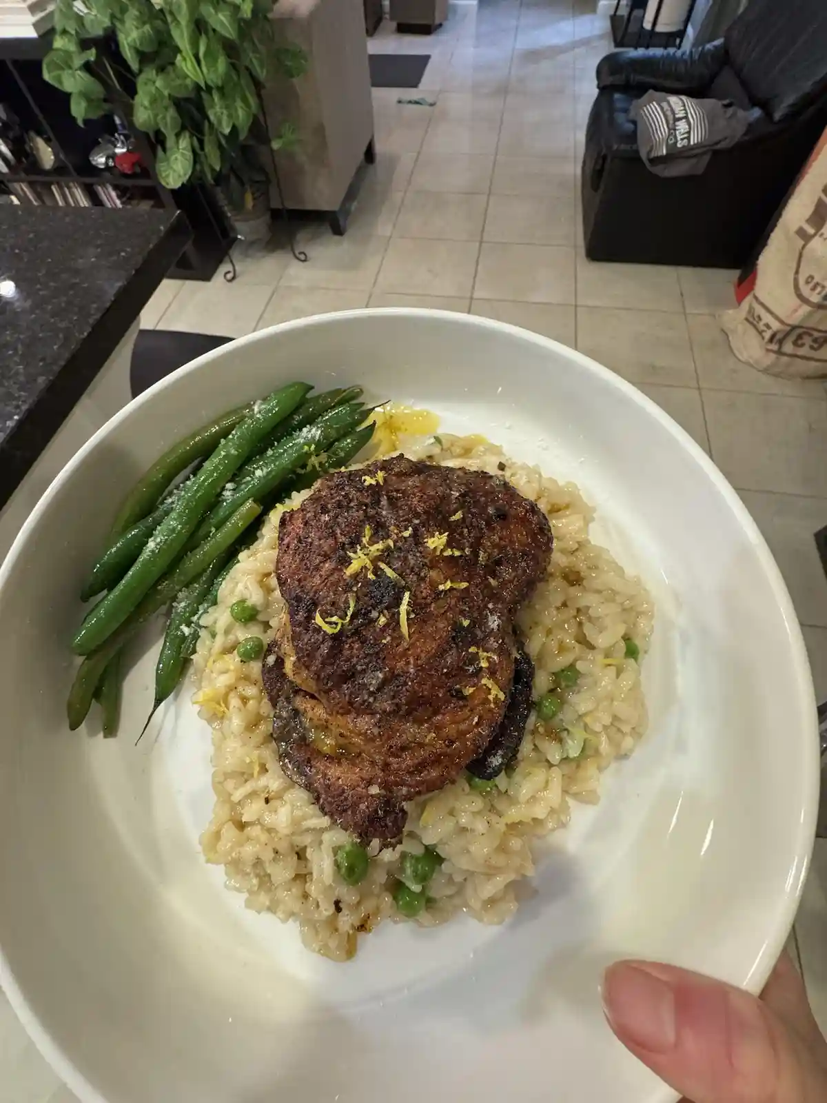 A person holds a white bowl containing a plated meal of risotto with peas, a seared meat patty, and green beans, garnished…