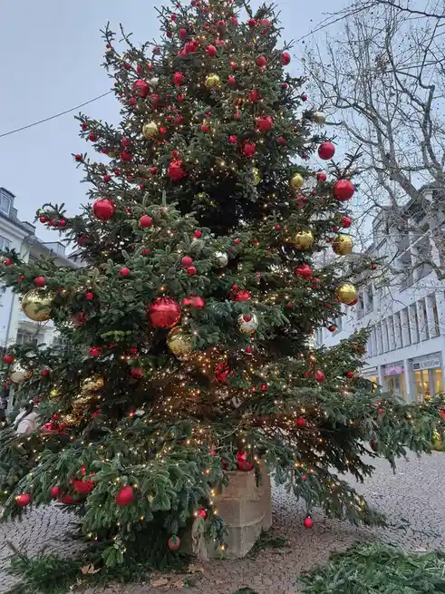 a large, heavily decorated christmas tree with red and gold ornaments and string lights is visibly leaning over to one side…