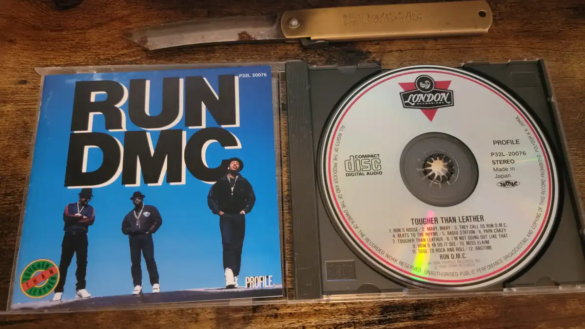 A CD case for Run-DMC's album 'Tougher Than Leather' is open, showing the cover art with the three band members and the…