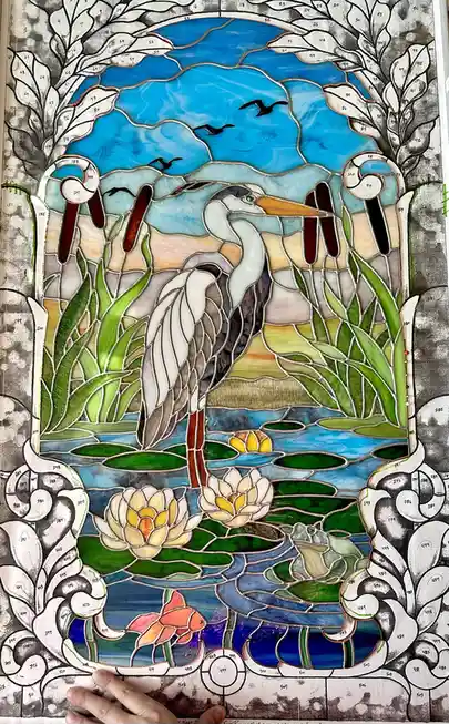 A completed stained glass artwork depicting a heron standing in a pond with lily pads and water lilies, surrounded by reeds…