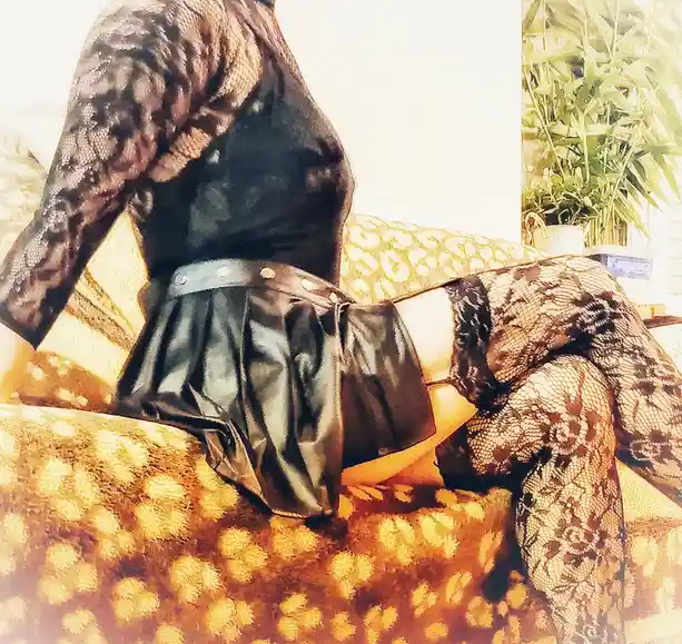 A person wearing black lace top and thigh-highs with a black leather skirt sits on a yellow patterned couch, viewed from…
