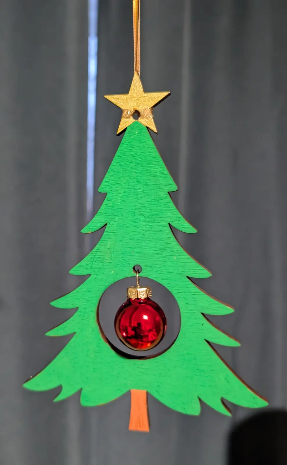 A green wooden cutout of a Christmas tree with a gold star on top and a red ornament hanging from a hole in the middle. The…