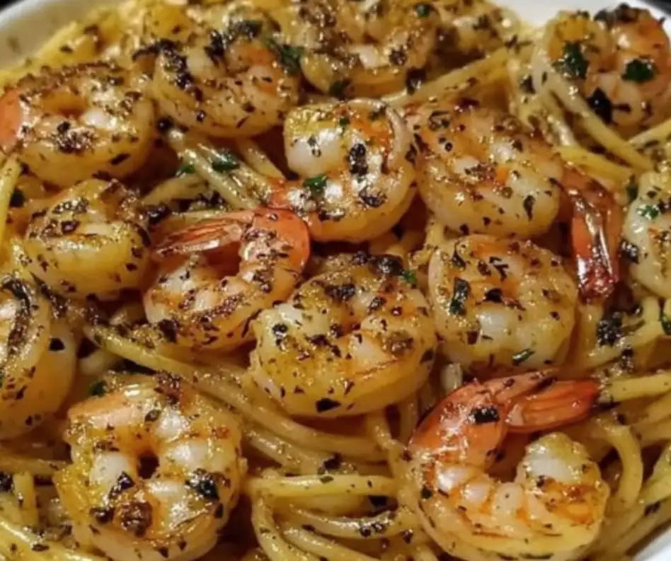 A close-up photo of a dish of spaghetti with large, cooked shrimp, both coated in a sauce and sprinkled with herbs and black…