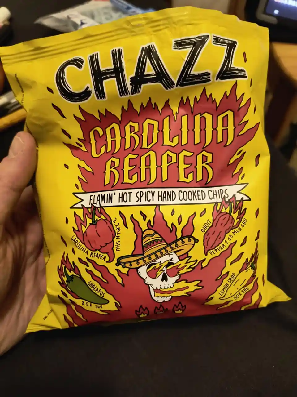 A hand holds a bright yellow bag of 'Chazz Carolina Reaper' chips, featuring fiery graphics with a skull wearing a sombrero…