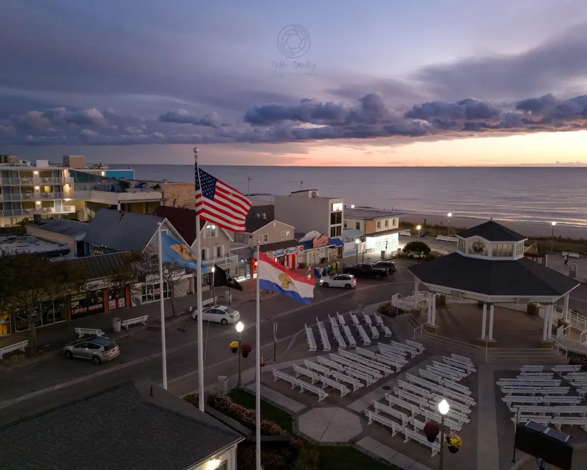 An aerial photo of a coastal town at dusk, showing rows of white benches set up for an event near a gazebo, with the…