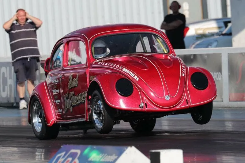 A red, modified Volkswagen Beetle race car with 'Bad Influence' and 'Wilkinson Brothers' written on its side is airborne on…