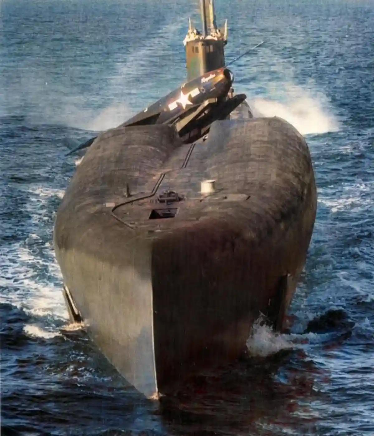 A military submarine is shown moving through ocean water, with a missile mounted on its deck. The missile has a white star…