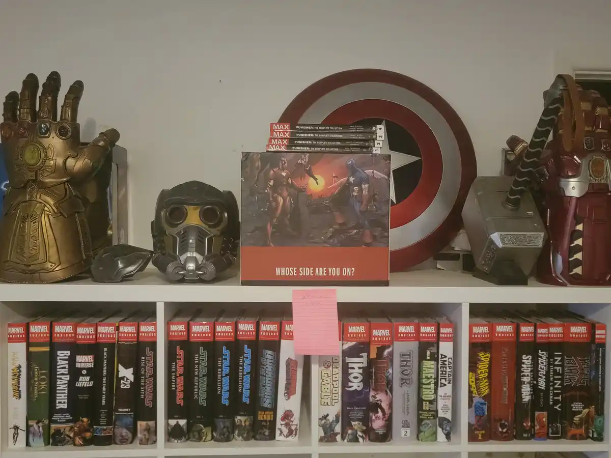 A bookshelf displays Marvel comic books and superhero memorabilia, including a Thanos gauntlet, a Gamora helmet, Captain…