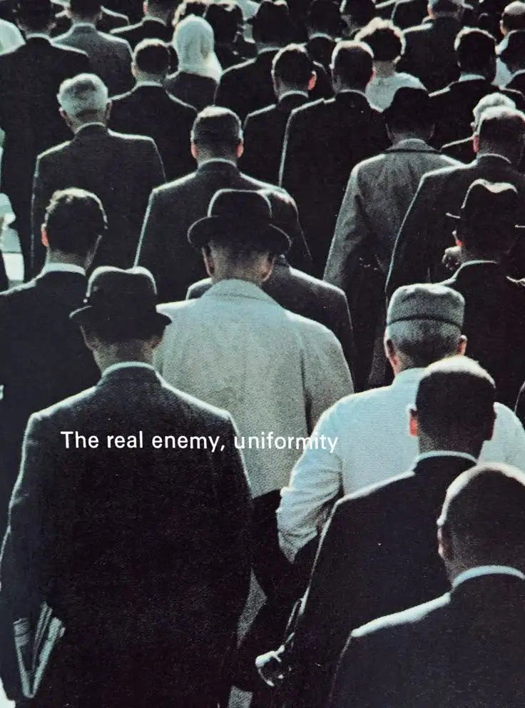 A vintage black-and-white photo shows a dense crowd of men viewed from behind, most wearing dark suits and hats, creating a…