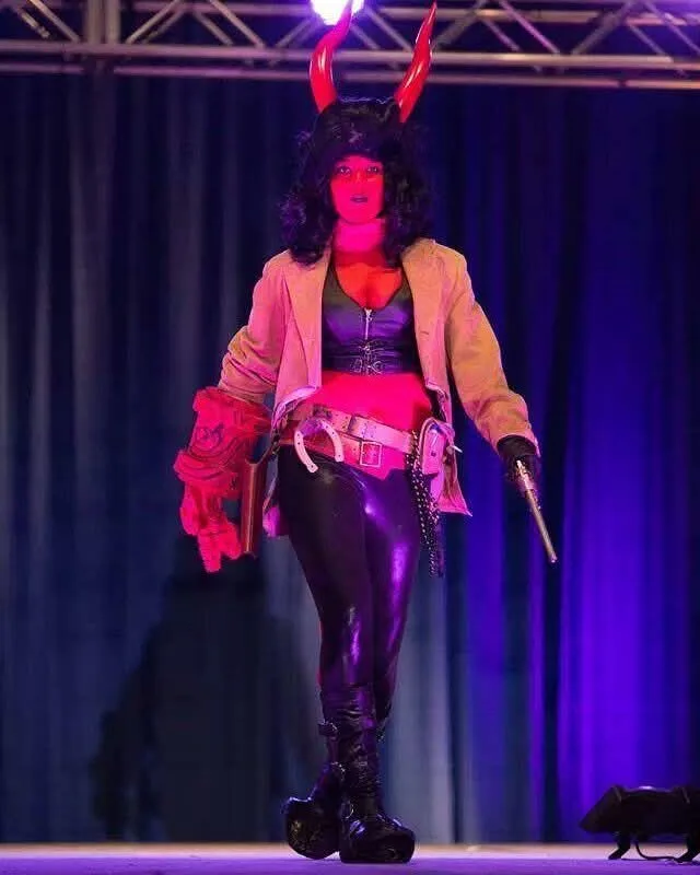 A person in a devil costume with red horns, black wig, leather pants, and a jacket walks on a stage under purple lighting…