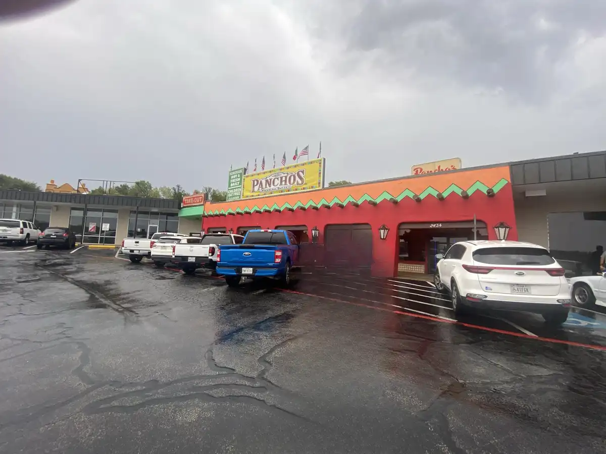 A wet parking lot outside a red and orange Pancho's Mexican Grill restaurant under a cloudy sky, with several cars parked…