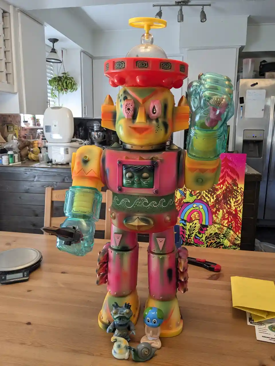 A large, colorful, spray-painted robot toy stands on a wooden kitchen table, holding a smartphone in one hand. Its chest…