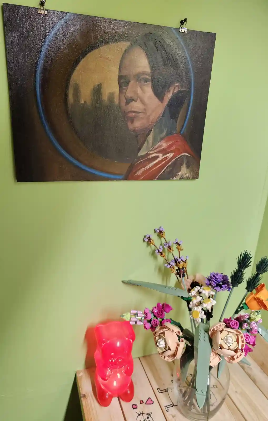 A painting of a person with a cityscape behind them hangs on a green wall, clipped with binder clips. Below, on a wooden…