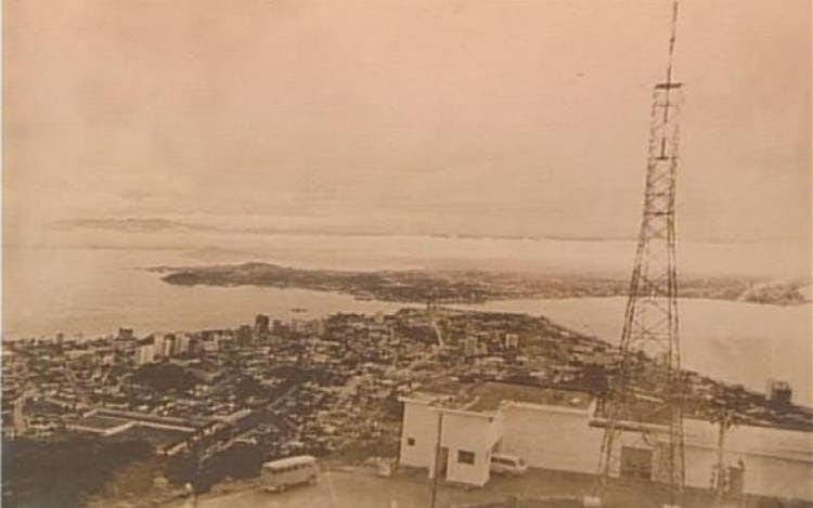 A sepia-toned, vintage aerial photograph shows a coastal city with a prominent, tall metal lattice tower on the right side…