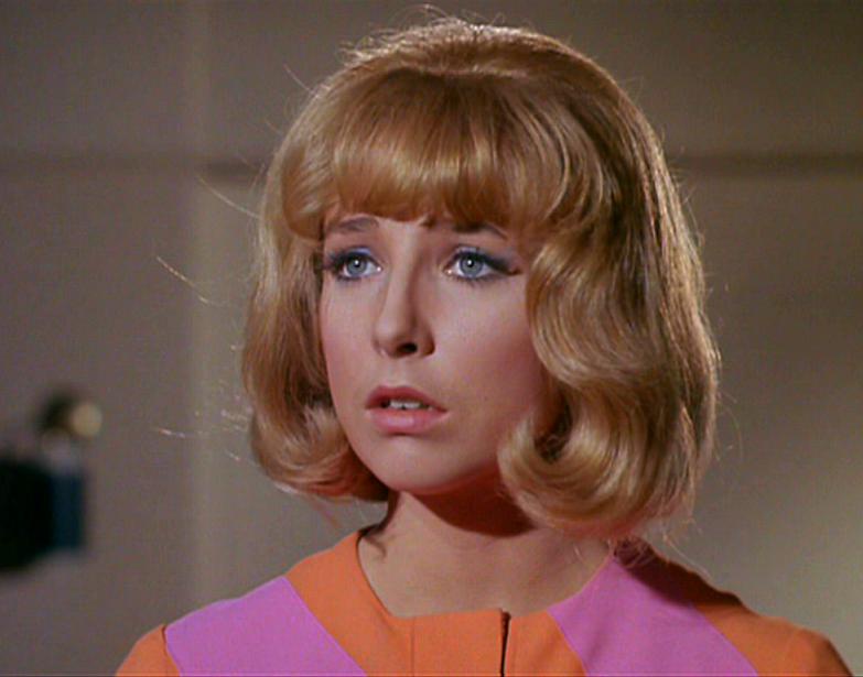 a woman with blonde bob hair and blue eyes wears a pink and orange outfit, looking shocked or dismayed. overlaid text reads…