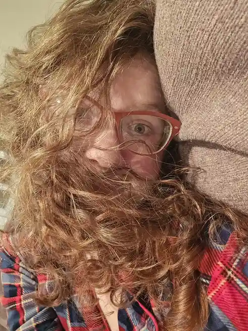 A person with curly reddish-brown hair and red-framed glasses is peeking out from behind a beige couch cushion, their face…