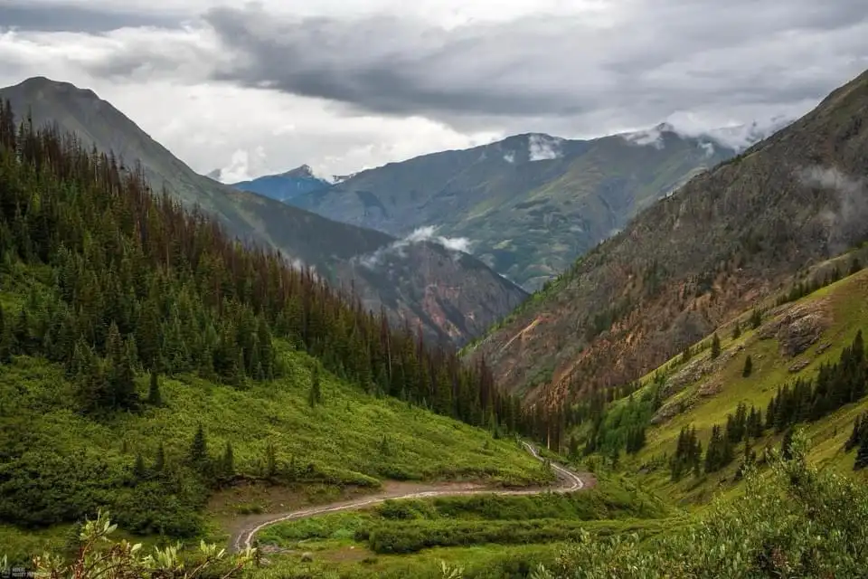 A scenic mountain valley with a winding dirt road cutting through lush green forests and rocky slopes under a cloudy sky…