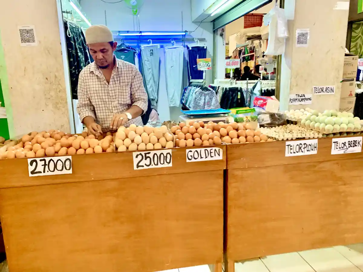 A man in a market stall arranges eggs behind a wooden counter labeled with prices and types like '27.000', '25.000 GOLDEN'…